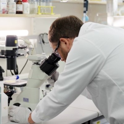 a man bends to look into a microscope at a container of yellow liquid watched on by another man, both are in white coats